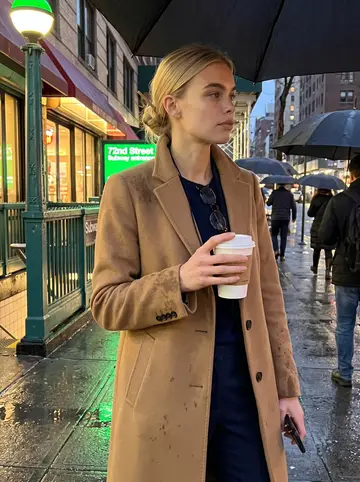 Fenna, AI character: outside the 72nd Street subway entrance after a late shift, wet pavement and umbrella tops around her