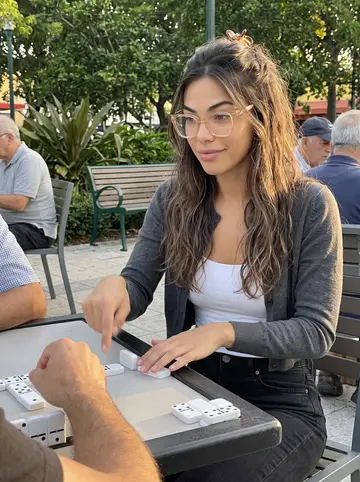Daniela, AI character: Domino Park on Calle Ocho in late afternoon, seated at an outdoor domino table with older men cropped…