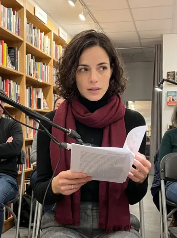 Lydia, AI character: a small bookstore reading night near the MFA program, shelves and folding chairs behind her