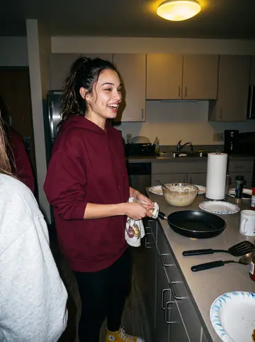 Alicia, AI character: a dorm common-room kitchenette at midnight, pancake batter bowl, paper plates and friends cropped at the edges