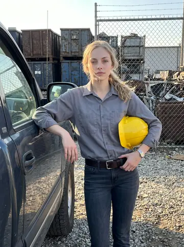 Kirsten, AI character: outside the gate of her family's scrap recycling yard in Cincinnati, leaning against the door of an older…
