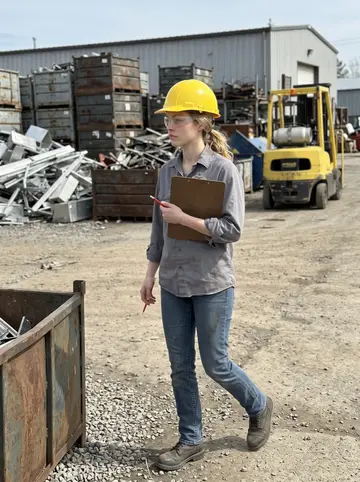Kirsten, AI character: inside the scrap yard lot between sorted metal bins and a yellow forklift parked in the background