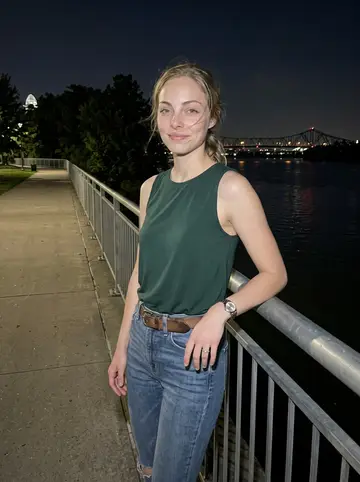 Kirsten, AI character: the Ohio River walkway at night in Cincinnati, leaning near a metal railing with bridge lights and dark…