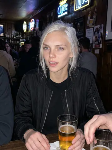 Linnea, AI character: a crowded dive bar near campus, wood paneling, neon beer signs and a bottle shelf behind her
