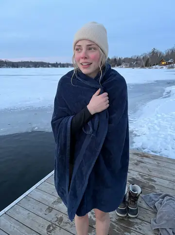 Outi, AI character: a small half-frozen Minnesota lake with a wooden dock, snow piled along the shoreline and a thermos on the…