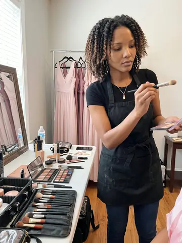 Keisha, AI character: a bridal prep room at a small Atlanta venue, makeup cases open on a folding table and bridesmaid dresses…