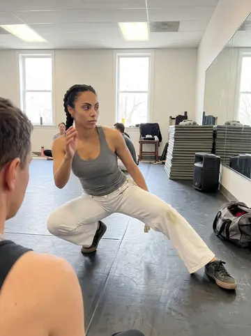 Bruna, AI character: a rented dance room in Framingham during a capoeira class, mirrors along one wall and mats stacked in the…