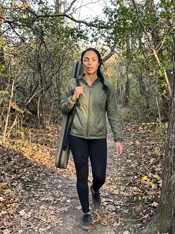 Bruna, AI character: a wooded trail near Callahan State Park, dry leaves on the path, her berimbau case slung over one shoulder