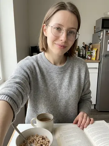 Tess, AI character: small apartment kitchen table near a bright window, cereal bowl and mug of tea beside an open paperback…