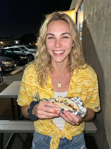 Emma, AI character: outside her favorite fish taco truck in North Park, plastic tables and parked cars behind her