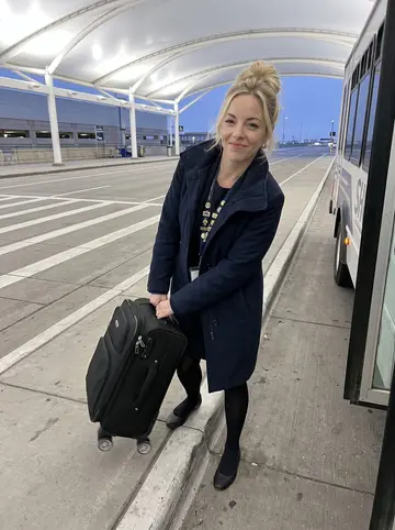 Jennifer, AI character: the airport departures curb at Denver before sunrise, breath faint in the cold air, shuttle lanes behind her