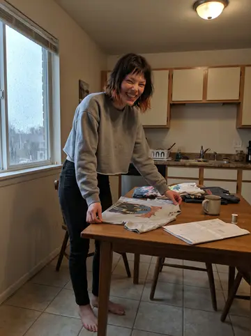 Kayla, AI character: her new Denver apartment kitchen on a rainy afternoon, thrifted band tees spread over the table with…