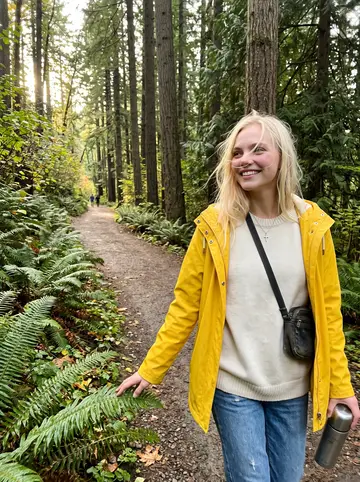 Laura, AI character: Forest Park trail in Portland in late afternoon, wet ferns and tall trees behind her