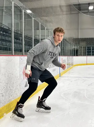 Gregory, AI character: inside the practice rink during a late team skate, boards and scuffed ice visible behind him