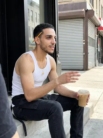 Yannis, AI character: the front stoop of a small shop on a Washington Heights block near 181st Street, a metal shutter and a…
