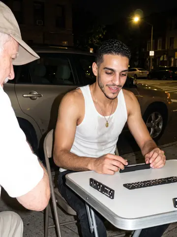 Yannis, AI character: a folding table of dominoes on the sidewalk of his block at night, plastic chairs, an old man's arm and…