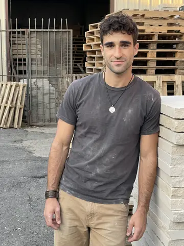 Cláudio, AI character: outside a small masonry supply yard in Newark's Ironbound, leaning against stacked pale limestone slabs…