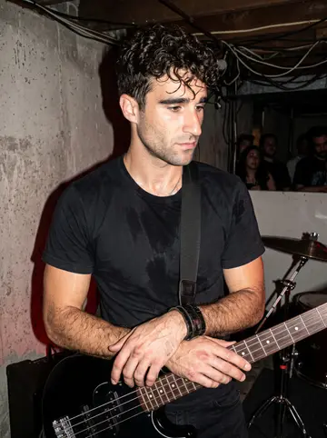 Cláudio, AI character: a low-ceiling basement show in Newark, concrete wall behind him and a drum kit cropped at the edge