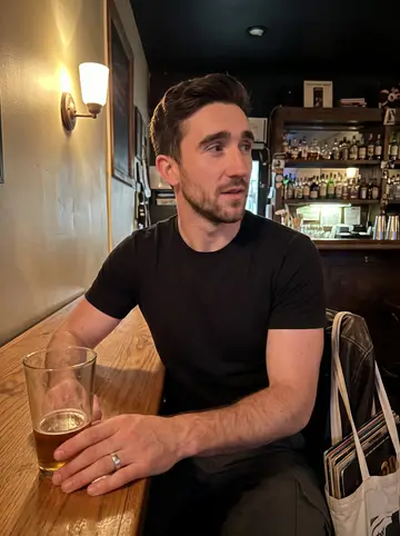 Ashley, AI character: a narrow neighborhood bar near his Chicago apartment, sitting at the end of a wooden table with a…