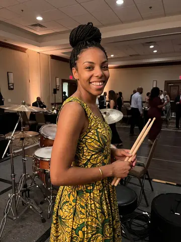 Onyinye, AI character: a Houston hotel ballroom during an Afrobeats wedding-band break, standing beside a drum kit near the edge…