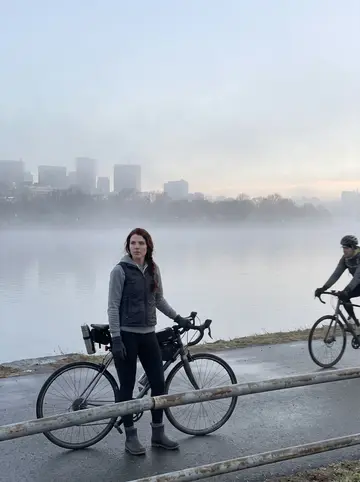 Donna, AI character: Charles River bike path before sunrise, thin fog over the water and Boston buildings still dim behind her
