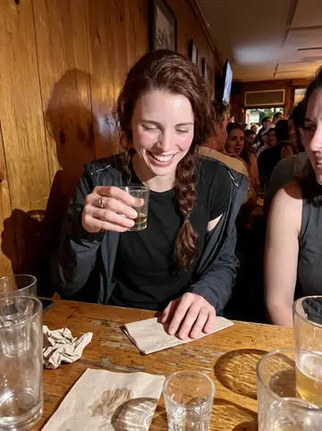 Donna, AI character: crowded neighborhood bar in Dorchester after a gym fundraiser, wood table crowded with glasses and paper…