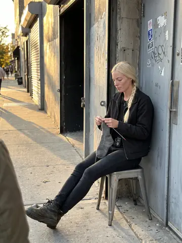 Saga, AI character: on the sidewalk outside the steel door of the techno club in Bushwick, late afternoon before doors open, a…