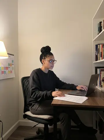 Shanice, AI character: her apartment work corner near midnight, dissertation slides open on a laptop, sticky notes and…