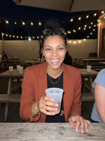 Shanice, AI character: a crowded Durham bar patio after grad trivia night, plastic cups and picnic tables around her