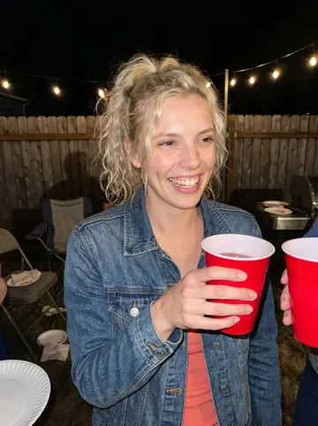 Amy, AI character: a crowded backyard birthday cookout in north Seattle after sunset, paper plates and patio chairs behind her