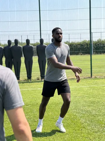 Obi, AI character: the grass practice field at the team facility during summer training camp, tall fence and a row of…