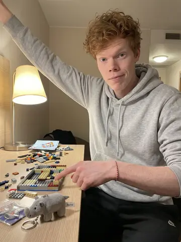 Patrick, AI character: team hotel desk on an away trip, half-built Lego set spread beside a packet of arena pins