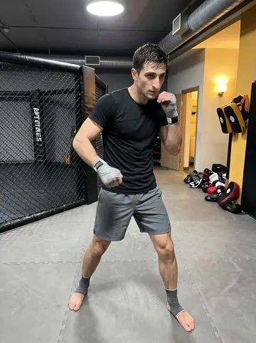 Jonny, AI character: inside the MMA gym near the cage, mats scuffed from training, gloves and pads stacked by the wall