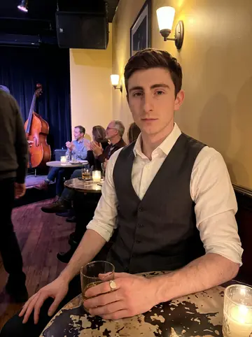 Conal, AI character: crowded Boston pub corner during a Celtic fiddle session, dark wood wall and small tables behind him