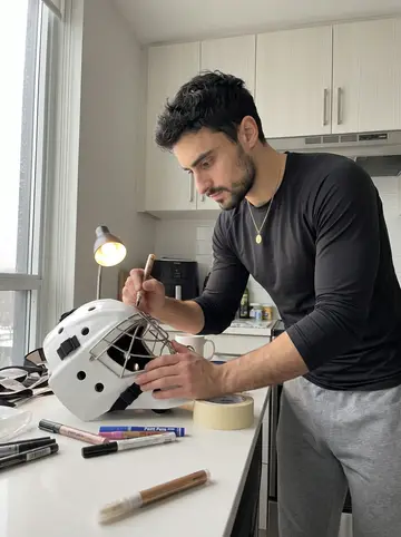 Modesto, AI character: his condo kitchen table on a rainy spring afternoon, spare goalie mask parts, paint pens, tape, and a mug…