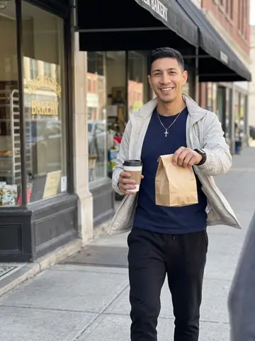 Yoandy, AI character: sidewalk outside a Rochester bakery on a bright spring afternoon, paper pastry bag in one hand and takeout…