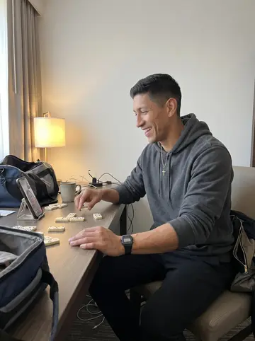 Yoandy, AI character: plain hotel room desk during an away trip, dominoes laid out beside a phone propped for a video call with…