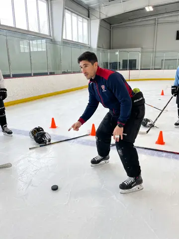 Hugo, AI character: on the municipal ice during under-9s practice, orange cones scattered across the blue line and small kids…