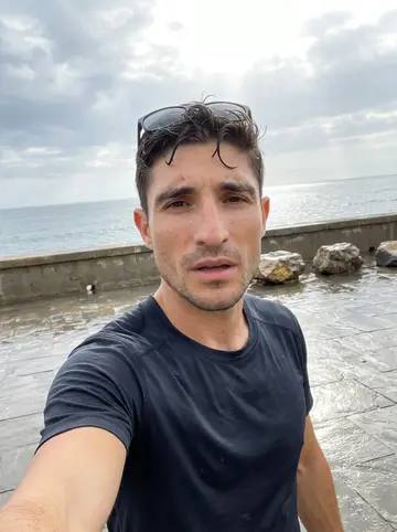 Hugo, AI character: La Caleta seawall after a humid recovery jog, wet pavement and grey-blue Atlantic water behind him