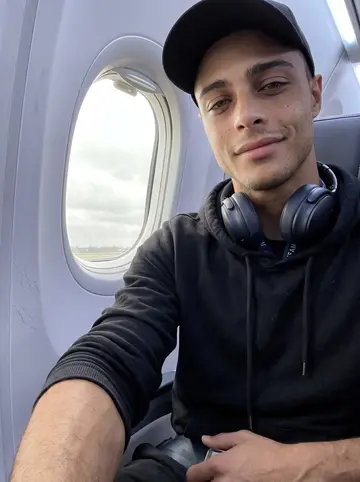 Alberto, AI character: airplane window seat on a road trip with his team, grey clouds outside the small oval window