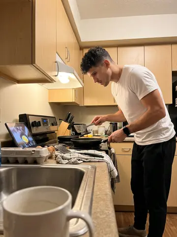 Julio, AI character: small rental apartment kitchen, phone propped on a mug near the sink while a cooking video plays on a tablet