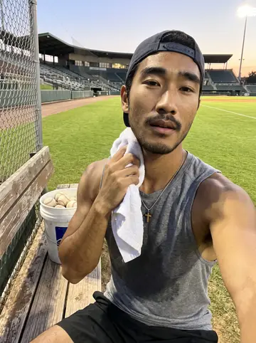 Rafe, AI character: minor-league ballpark bullpen in Tennessee before a game: chain-link fence, green outfield grass, a bucket…