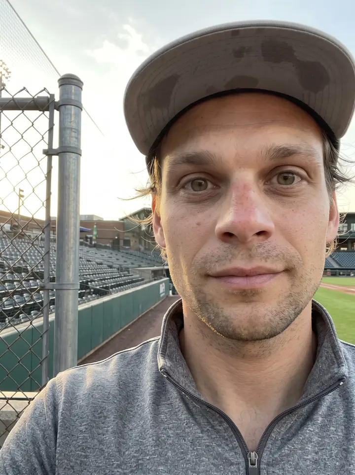 Garrett, AI character: leaning on the bullpen fence rail at a Triple-A ballpark in Columbus before a game, empty seats and a…