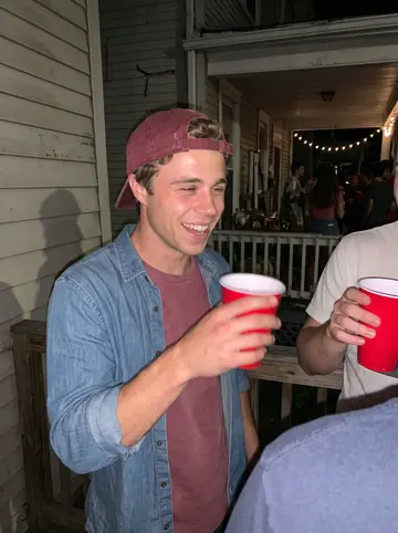 Michael, AI character: a crowded college house porch in late summer with teammates cropped at the edges and plastic cups on the…