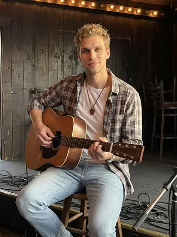 Andrew, AI character: a small Lower Broadway bar stage before the doors open, dark wood wall behind him, a mic stand and his…