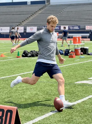 Andrew, AI character: outdoor football practice field in Nashville, green turf, empty lower stands and sideline gear behind him