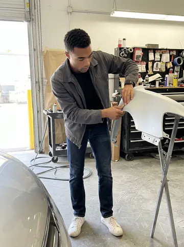 Prince, AI character: inside the body shop bay during a busy noon, with a primed fender on a stand and a toolbox wall behind him