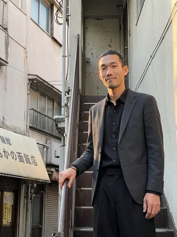 Ryo, AI character: a narrow Koenji side street below his real estate office, the noodle shop awning behind him and late sun…