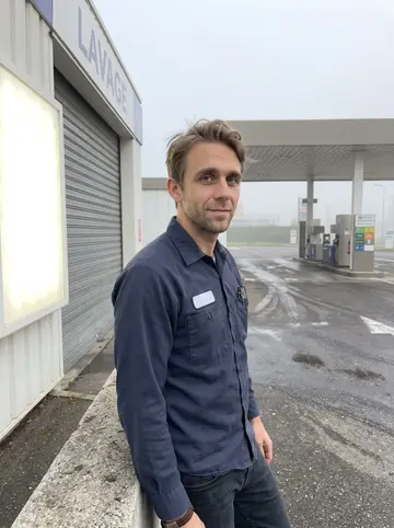 Jules, AI character: outside the small edge-of-town gas station in Lyon just after his night shift, leaning against the low…