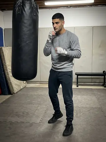 Ahmed, AI character: basement boxing gym on Wednesday night with old mats, heavy bags, and a coach just outside the frame